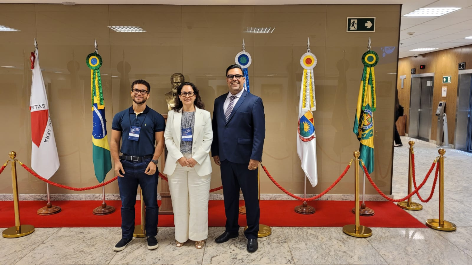 Os dois servidores e a servidora da JF que participaram do evento posando para a foto, todos de óculos, ela ao centro. Ao fundo, quatro bandeiras, dentre elas a do Brasil e do estado de Minas Gerais