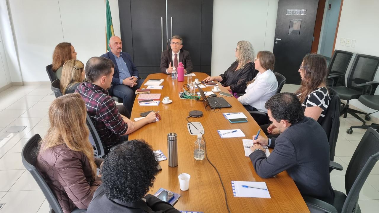 Um grupo de pessoas está reunido ao redor de uma mesa retangular de madeira em uma sala de reuniões. Sobre a mesa há diversos itens: cadernos, canetas, copos, uma garrafa de água, um notebook, um recipiente de vidro com água, cabos e uma garrafa térmica rosa. A sala tem paredes brancas e piso de cerâmica clara. Ao fundo, há um armário preto com duas portas, e ao lado dele, uma bandeira do Brasil. As pessoas estão envolvidas em uma conversa, algumas tomando notas e outras ouvindo com atenção.