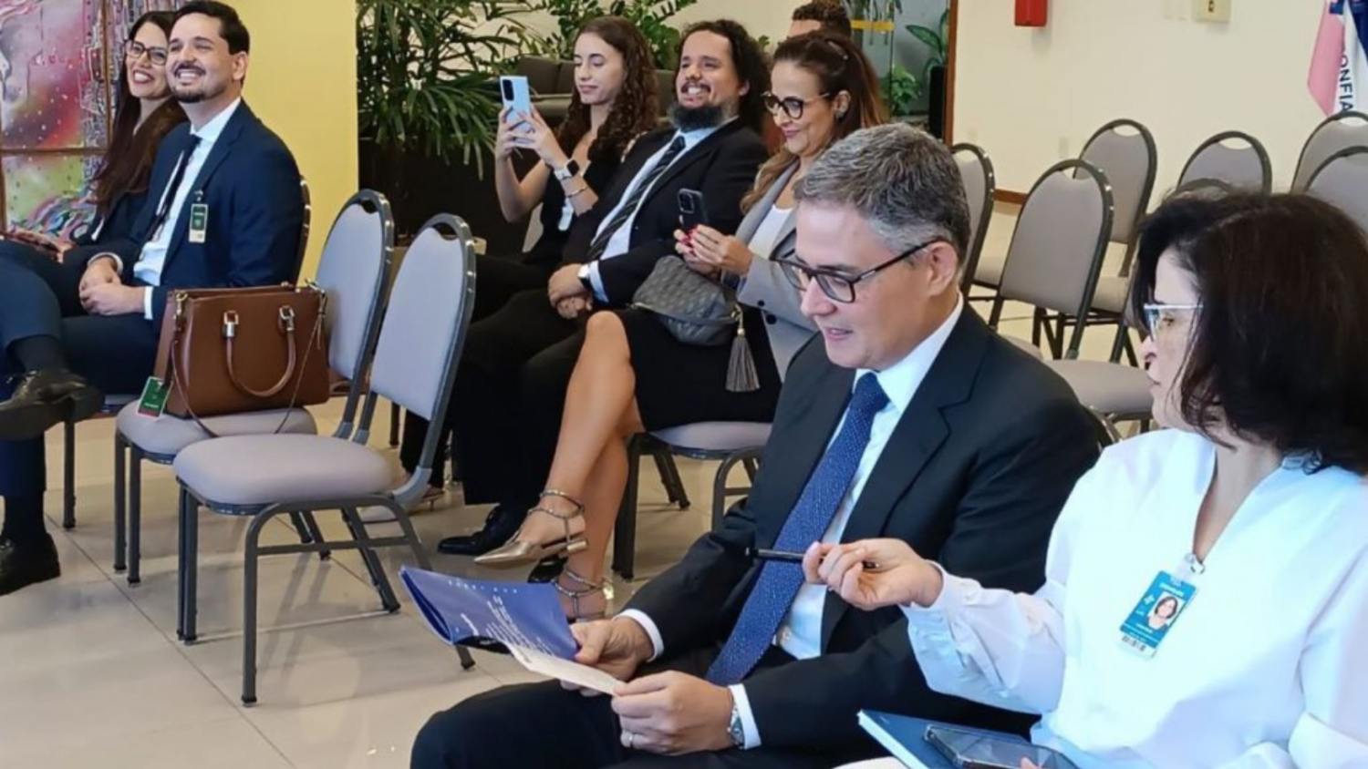 imagem de magistrados, servidores e professores sentados assistindo à cerimônia. Nela aparece o diretor do foro folheando o guia e a diretora da secretaria geral apontando algo no manual