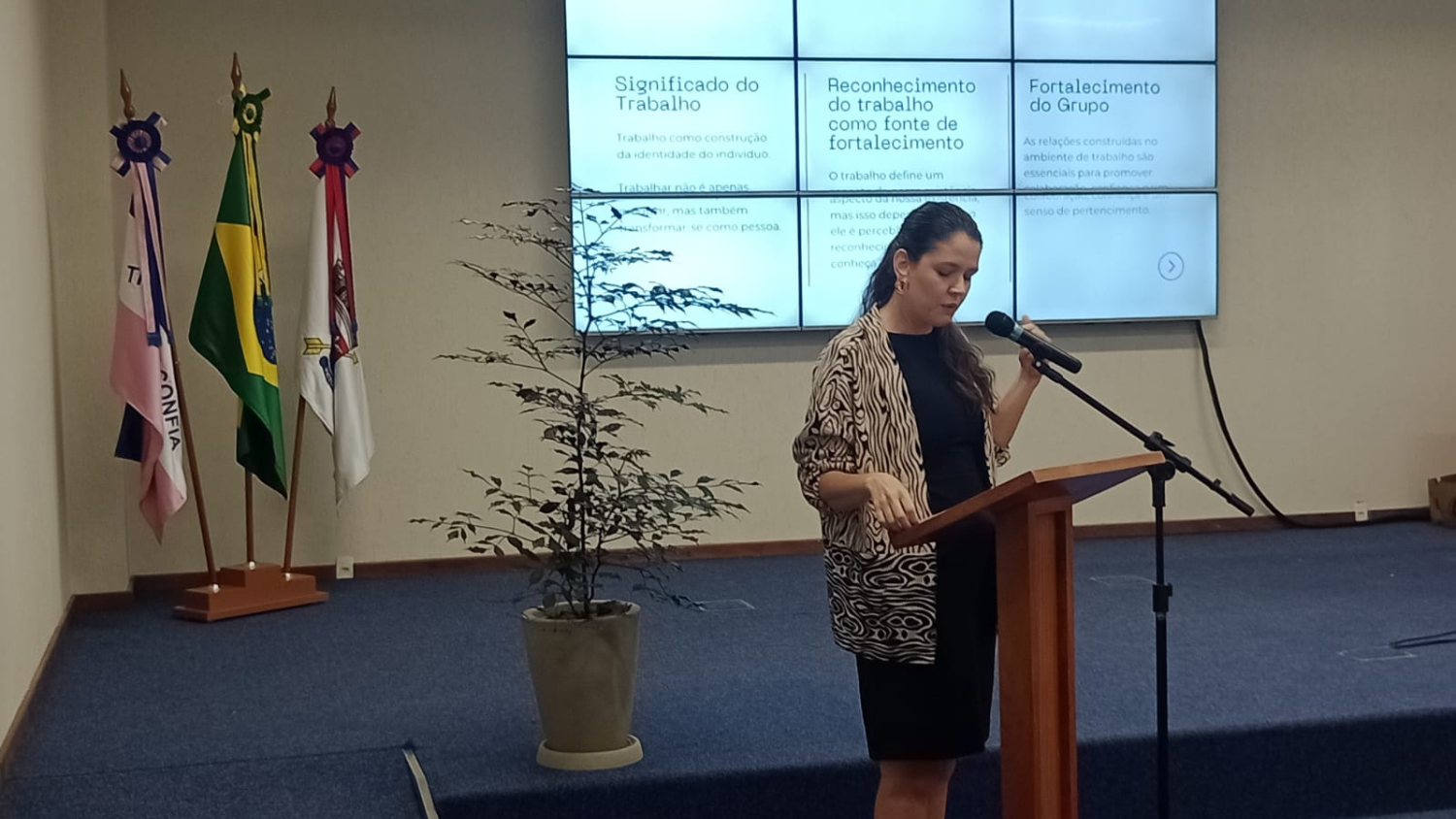 Mulher branca, de rabo de cavalo, com vestido preto e cardigã com estampa em branco e marrom, fala de pé ao microfone