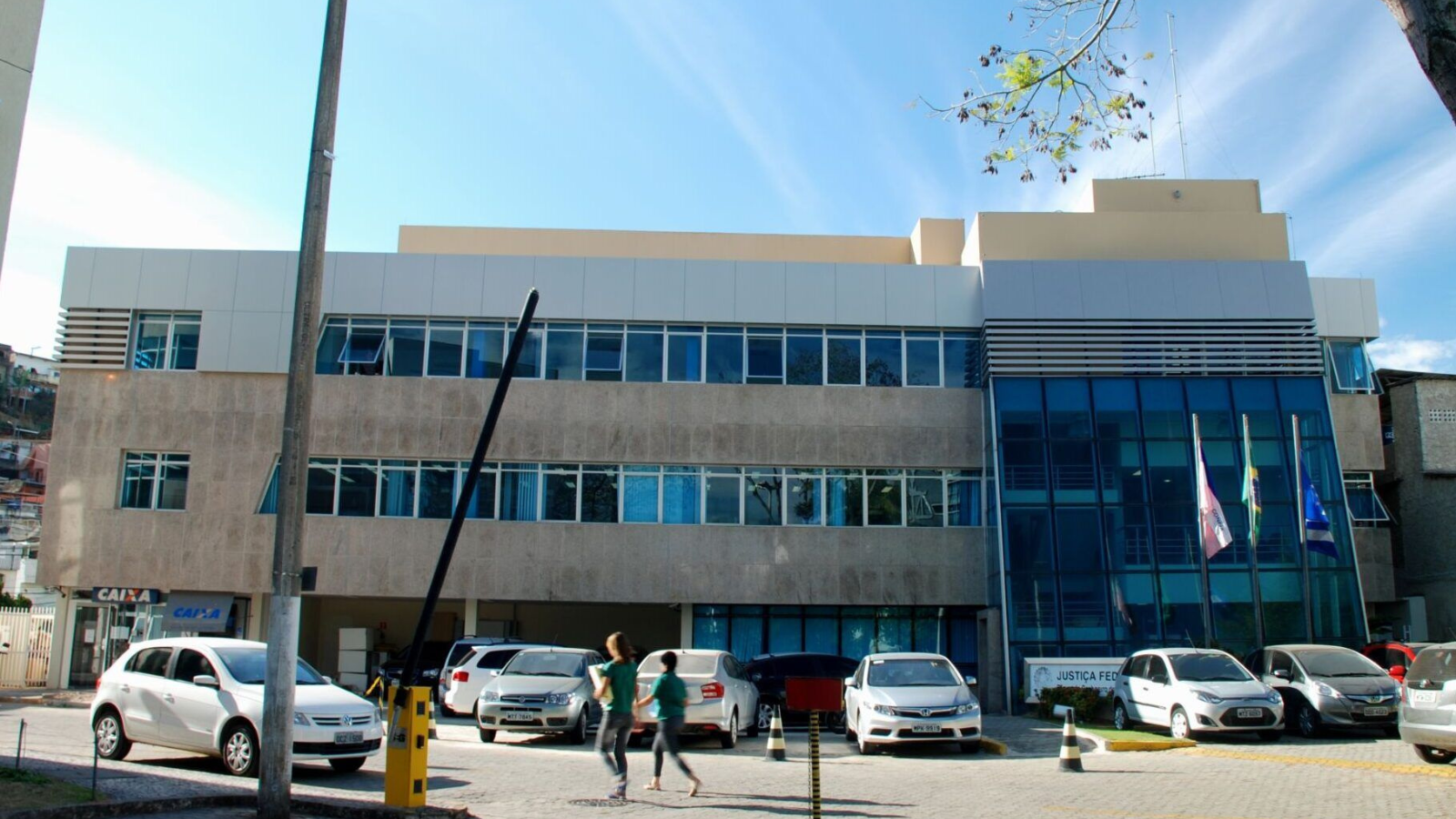 fachada de prédio horizontal com dois andares, carros estacionados em frente, e três bandeiras hasteadas: do Espírito Santo, do Brasil e de Cachoeiroo