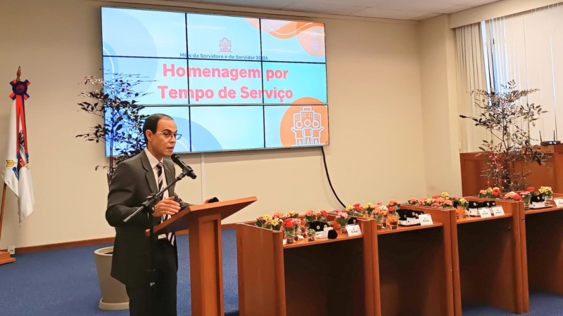Um homem de terno cinza, camisa branca e gravata fala ao microfone. Ao seu lado mesas de madeira com flores coloridas e bótons com brasão da República. Ao fundo, telão na parede com imagem abstrata em tons de azul e laranja e título: "Homenagem por Tempo de Serviço"