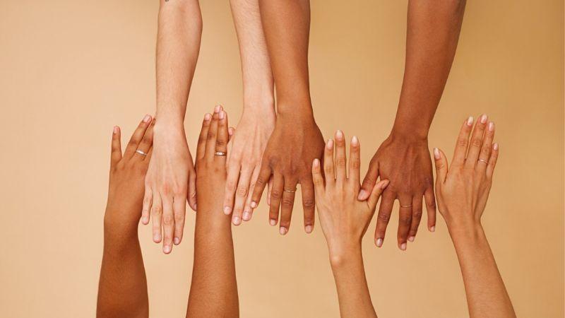 Foto de 8 mãos de pessoas brancas, pardas e pretas, uma do lado da outra, sobre um fundo bege