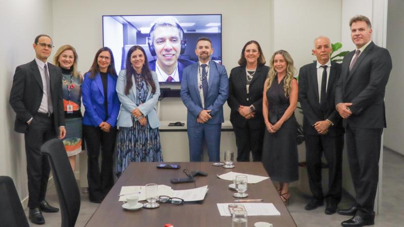 4 homens e 5 mulheres de pé, de frente para a câmera. No centro outro homem aparece num telão. À frente deles uma mesa retangular grande de madeira escura