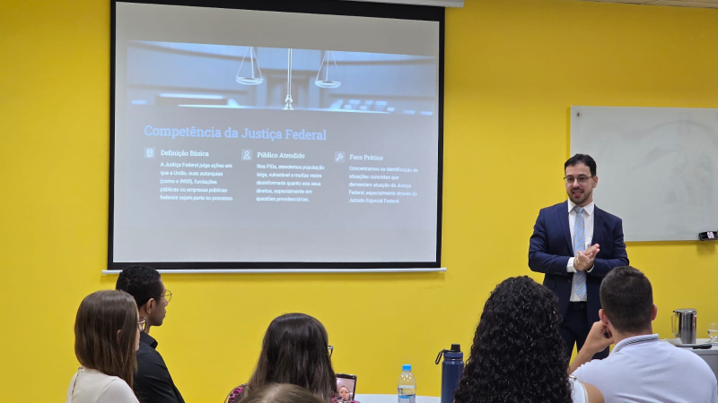 O juiz de pé dando sua aula. Ele veste terno escuro, camisa branca e gravata azul claro. Ao fundo, parede amarela e telão de LED