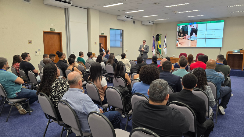 O juiz federal Ronald Krüger Rodor fala em pé no auditório. Ele está em frente a um telão com slides e, ao fundo, aparecem as bandeiras do Brasil e do Espírito Santo