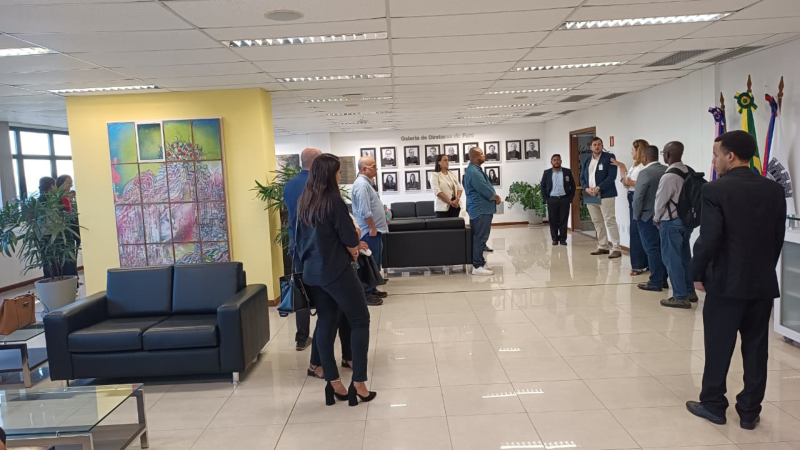 Estudantes e servidores visitam a sala de reuniões dos juízes federais, onde há a galeria de ex-diretores do foro da JFES. Eles estão organizados em semicírculo, ouvindo a explicação de uma servidora