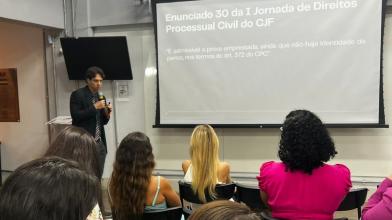 Juiz federal Caio Souto Araújo apresenta palestra segurando microfone, diante de um telão que exibe o Enunciado 30 da I Jornada de Direitos Processuais Civis do CJF. O público assiste sentado