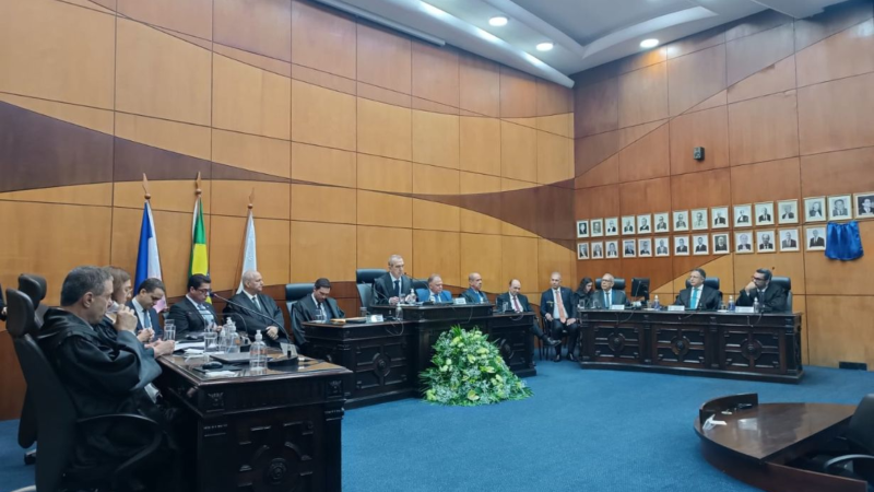 Solenidade de posse no plenário do TRE-ES, com magistrados e autoridades sentados à mesa de honra. Ao centro, o novo presidente conduz a sessão. Há um arranjo de flores no piso, e bandeiras ao fundo.