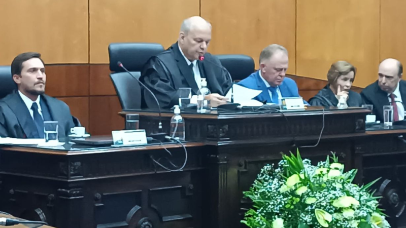 Mesa de honra durante a solenidade no TRE-ES. Um magistrado, ao centro, lê um documento ao microfone. Autoridades estão sentadas ao lado dele. Em primeiro plano, há um arranjo de flores verdes e brancas.