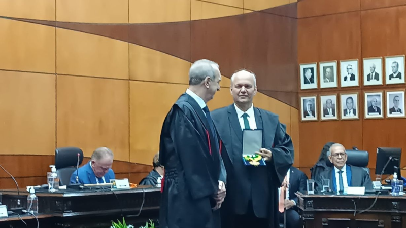 Momento da solenidade no TRE-ES em que um magistrado entrega uma honraria a outro. Ambos estão de toga, em pé, na frente da mesa de autoridades.