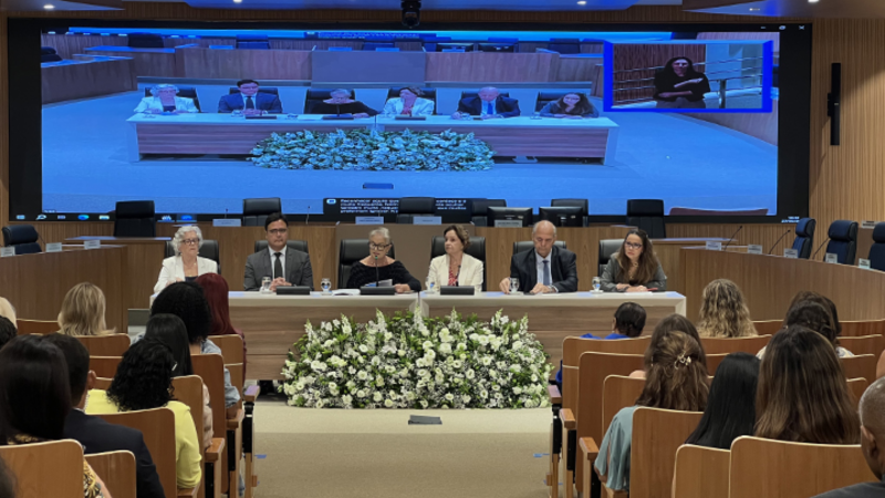 Auditório do Tribunal de Justiça lotado durante seminário sobre os desafios enfrentados pelas mulheres no Sistema de Justiça. À frente, seis integrantes compõem a mesa de abertura. Ao fundo, um telão exibe a transmissão do evento, com intérprete de Libras em destaque no canto superior direito. Há arranjo de flores brancas em frente à mesa.