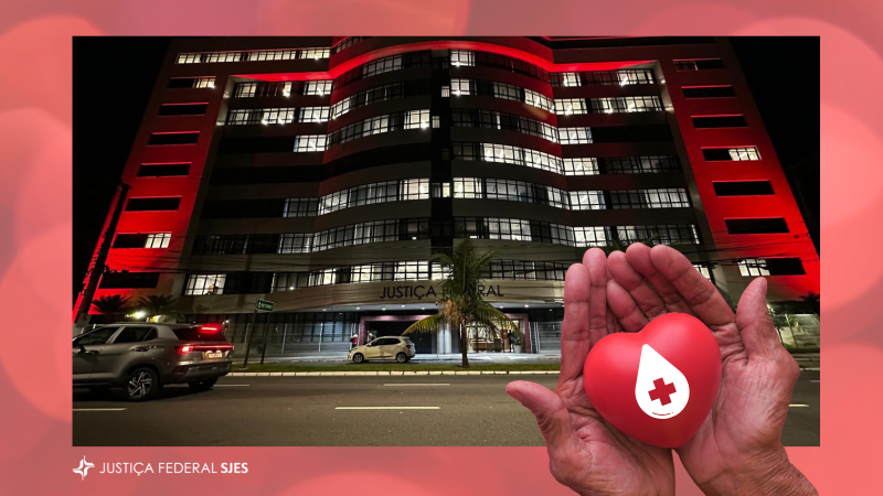 Fotografia noturna do edifício-sede da JFES iluminado em vermelho, em referência à campanha Junho Vermelho. No canto inferior direito da imagem, mãos em concha seguram um coração vermelho com o símbolo de uma gota de sangue e uma cruz branca