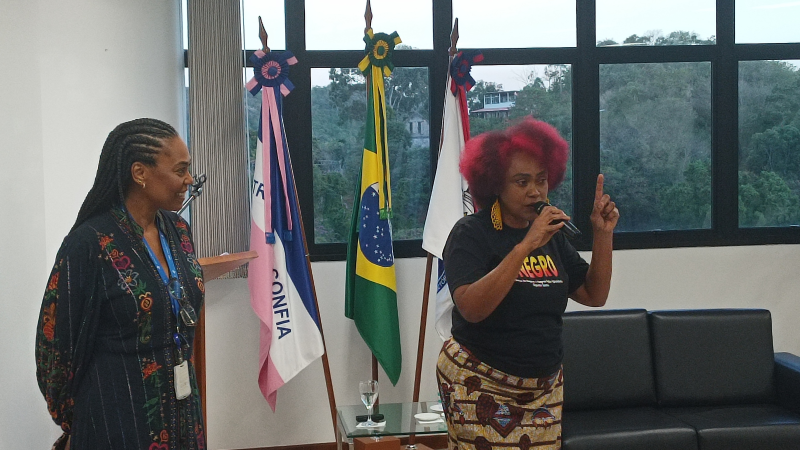 Duas mulheres pretas de pé. A da direita fala ao microfone e está com o dedo em riste. A da esquerda olha para ela sorrindo.