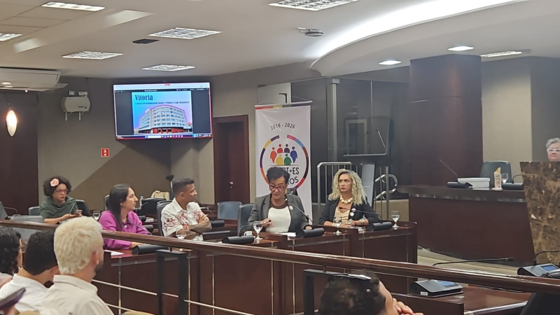 Cinco pessoas se apresentam sentadas em uma mesa, à frente do auditório. Uma delas lê um documento em voz alta