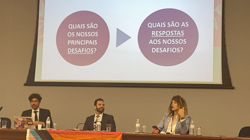 Mesa de debates com dois homens e uma mulher. A mulher fala. Ao fundo, telão com dois círculos na cor marrom e, sobre eles, texto na cor branca, com duas perguntas: Quais são os nossos principais desafios? Quais são as respostas aos nossos desafios?