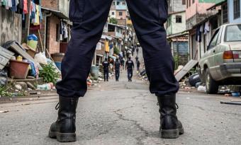 A cena mostra um grupo de policiais caminhando por uma rua estreita. O ponto de vista é de trás de um dos agentes em primeiro plano, destacando suas botas e pernas. Mais à frente, outros agentes aparecem distribuídos pela rua. O ambiente ao redor inclui casas pequenas e próximas umas das outras, roupas penduradas em varais, lixo espalhado pelo chão e um carro estacionado. O cenário transmite a ideia de uma operação policial em uma comunidade.