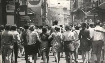 Fotografia em preto e branco de uma rua movimentada no centro urbano, com muitas pessoas caminhando juntas, vistas de costas. Há prédios altos, placas comerciais e uma multidão ocupando a via, sugerindo uma manifestação