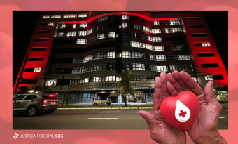 Fotografia noturna do edifício-sede da JFES iluminado em vermelho, em referência à campanha Junho Vermelho. No canto inferior direito da imagem, mãos em concha seguram um coração vermelho com o símbolo de uma gota de sangue e uma cruz branca
