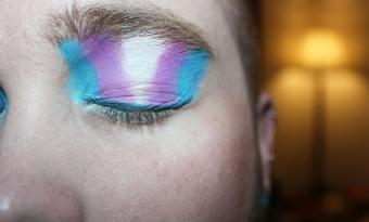 Close do rosto de uma pessoa com o olho fechado, usando maquiagem artística em tons de azul, rosa e branco, aplicada também sobre a sobrancelha. Ao fundo, desfocado, aparece um abajur aceso em ambiente interno com iluminação quente.