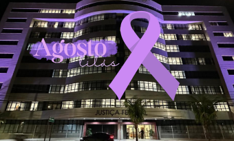 Fachada da sede da Justiça Federal, em Vitória, iluminada de lilás. Por cima da foto, um laço lilás e o texto "Agosto Lilás"