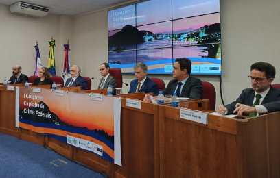 Mesa diretora, composta por sete autoridades (sendo seis homens e uma mulher). Ao fundo, na parede, telas de monitores com a imagem de paisagem da baía de Vitória no pôr do sol