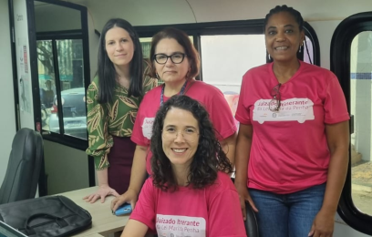 Foto de quatro mulheres (três em pé e uma sentada utilizando um notebook). Três delas usam uma camisa rosa, com estampa de letras brancas