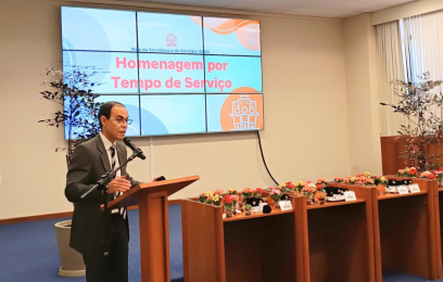 Um homem de terno cinza, camisa branca e gravata fala ao microfone. Ao seu lado mesas de madeira com flores coloridas e bótons com brasão da República. Ao fundo, telão na parede com imagem abstrata em tons de azul e laranja e título: "Homenagem por Tempo de Serviço"