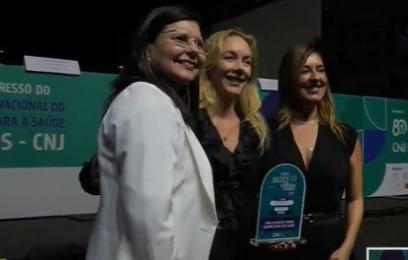 Três mulheres diante de uma bancada. A do meio segura o troféu