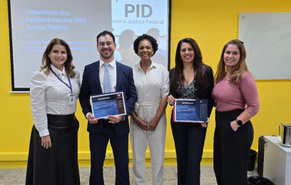 Cinco pessoas de pé, sorrindo para a foto: o juiz, a juíza, duas servidoras da Justiça Federal e uma instrutora da EMES. O juiz e a juíza seguram o certificado de participação. Ao fundo, parede amarela com tela de LED