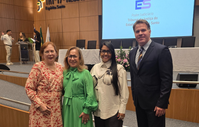 Quatro pessoas estão em pé e sorrindo para a foto, no auditório do Tribunal Regional do Trabalho da 17ª Região, no Espírito Santo. Ao fundo, há um telão com o logotipo do Tribunal de Justiça do Espírito Santo (TJES) e uma mesa com cadeiras vazias. Duas mulheres usam vestidos (um verde e um estampado em vermelho e branco), uma terceira mulher veste camisa branca com estampa preta e calça escura, e um homem está de terno e gravata.