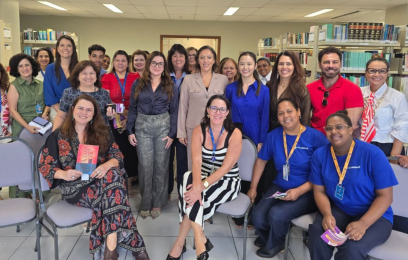 Grupo de cerca de 20 pessoas, em sua maioria mulheres, posando em pé e sentadas em uma biblioteca institucional, com estantes de livros ao fundo. Algumas seguram exemplares de livro. O ambiente é iluminado e formal, com clima de integração após atividade coletiva.
