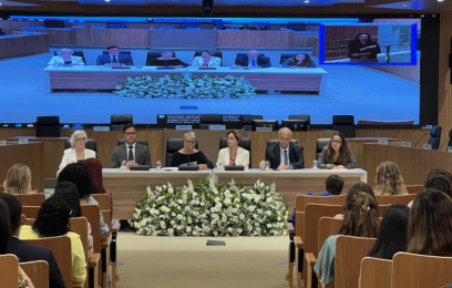 Auditório do Tribunal de Justiça lotado durante seminário sobre os desafios enfrentados pelas mulheres no Sistema de Justiça. À frente, seis integrantes compõem a mesa de abertura. Ao fundo, um telão exibe a transmissão do evento, com intérprete de Libras em destaque no canto superior direito. Há arranjo de flores brancas em frente à mesa.