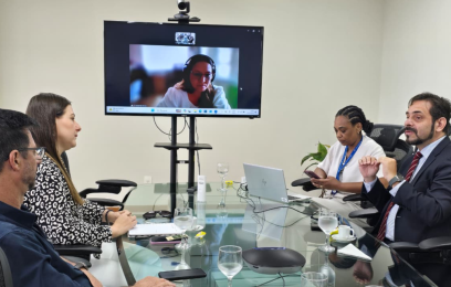A imagem mostra uma reunião em uma sala: quatro pessoas estão sentadas ao redor de uma mesa de vidro, enquanto uma participante aparece em uma tela ao fundo, em videoconferência