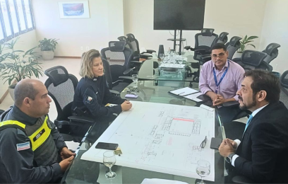 Quatro pessoas participam de uma reunião em uma sala de conferências, sentadas ao redor de uma mesa de vidro com uma planta aberta sobre ela. Entre os participantes estão um policial militar, uma guarda municipal e dois representantes da Justiça Federal. Ao fundo, há cadeiras, plantas e uma televisão.