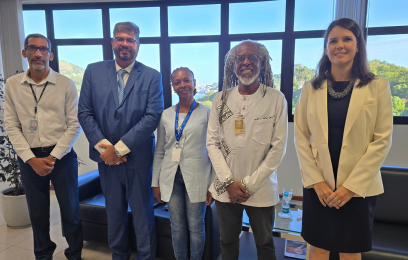 Cinco pessoas posam para foto em pé, lado a lado, em ambiente interno iluminado por grandes janelas. Entre elas está o professor Gustavo Forde, sorrindo, com cabelos em dreadlocks e roupa clara. Os demais participantes vestem trajes formais ou institucionais, incluindo terno e crachá. Ao fundo, vista externa com vegetação e água.