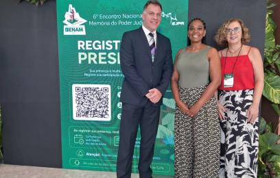 Um homem e duas mulheres, de pé, sorrindo para a câmera. Ao fundo, banner verde com letras brancas, com informações sobre o evento. Plantas nas laterais enfeitam o ambiente