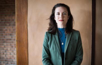 Mulher branca, cabelos pretos até o ombro, veste blazer verde. 