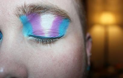 Close do rosto de uma pessoa com o olho fechado, usando maquiagem artística em tons de azul, rosa e branco, aplicada também sobre a sobrancelha. Ao fundo, desfocado, aparece um abajur aceso em ambiente interno com iluminação quente.