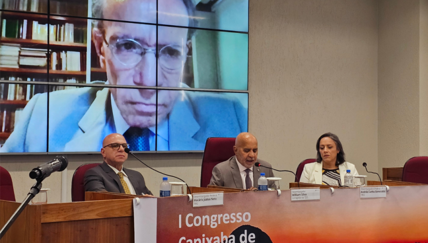 Dois homens e uma mulher sentados em mesas vermelhas, por trás de módulos de madeira, de frente para o auditório. O homem do meio fala ao microfone. Ao fundo, telão com a imagem de um homem